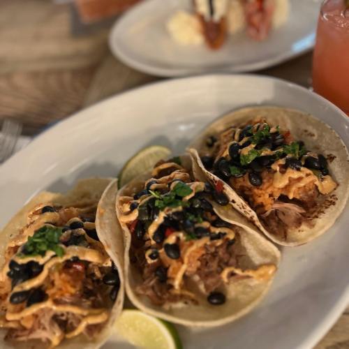 Three tacos filled with pulled pork, black beans, and drizzled sauce, served with lime wedges on a white plate.