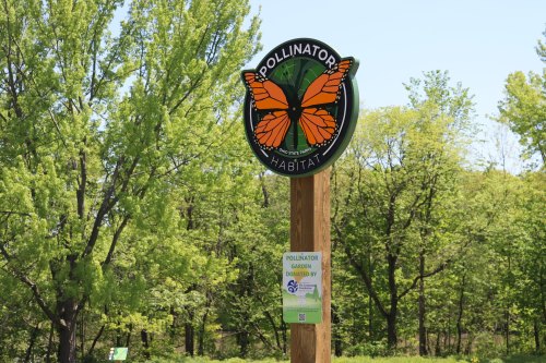 A sign for a pollinator habitat featuring an orange butterfly, surrounded by green trees and nature.