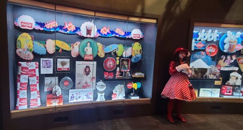 A display case filled with vintage Coca-Cola memorabilia, featuring colorful items and a woman in a polka dot dress nearby.
