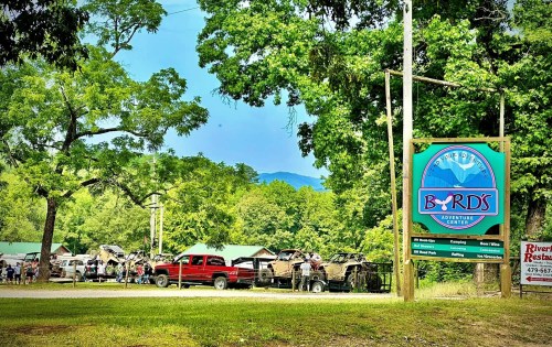 A sign for Byrd's Adventure Center surrounded by trees, with parked vehicles and mountains in the background.