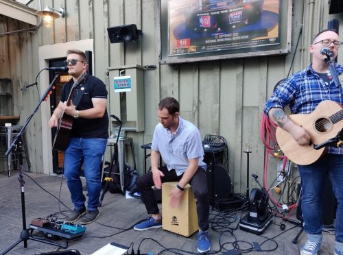 Three musicians perform live outdoors, with one on guitar, another on a cajón, and the third playing an acoustic guitar.