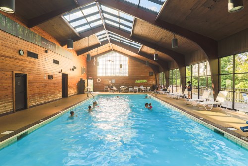 Indoor swimming pool with wooden walls, sunlight streaming through large windows, and people swimming and relaxing.
