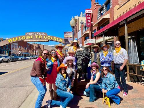A group of people in cowboy attire poses by a bison statue under a "Welcome to Golden" sign on a sunny day.
