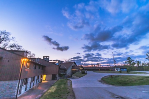 A building at dusk with a cloudy sky, featuring a well-lit entrance and a winding driveway.