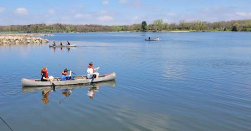 Two people in a canoe fishing on a calm lake, with other boats and greenery in the background under a clear blue sky.