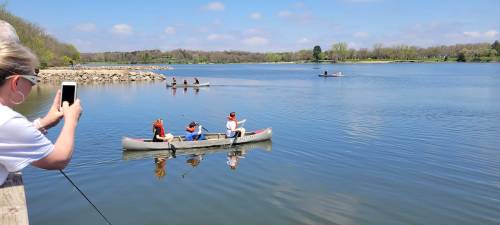 A person takes a photo while children paddle a canoe on a calm lake, with others rowing in the background.