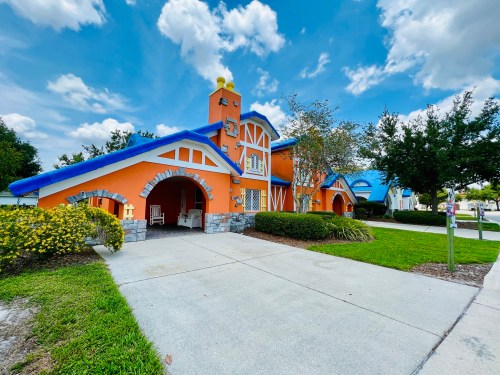 Colorful house with orange walls, blue roof, and stone accents, surrounded by greenery and a clear blue sky.