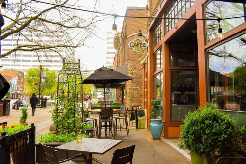 Outdoor café scene with tables, umbrellas, and greenery, set against a brick building in an urban area.