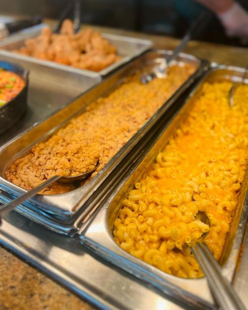 A buffet display featuring trays of macaroni and cheese, fried chicken, and a colorful vegetable mix.