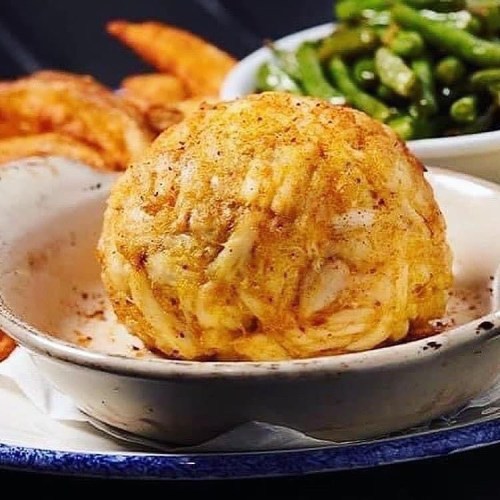 A golden, crispy crab cake served in a dish, accompanied by green beans and shrimp in the background.