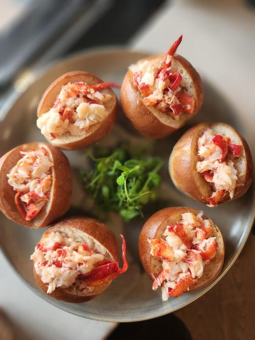 A plate of six lobster rolls in soft buns, garnished with fresh greens.
