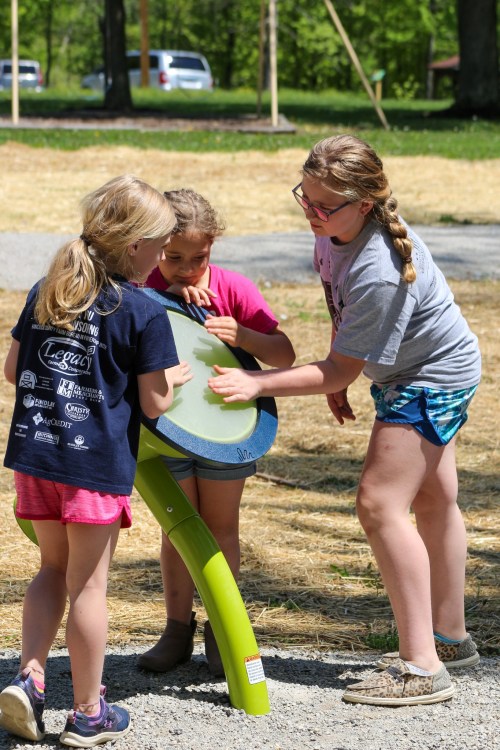 Three children interact with a green playground equipment piece in a sunny outdoor setting.
