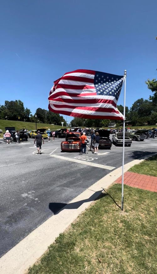 A U.S. flag waves in the foreground as people gather around cars at a sunny outdoor event.