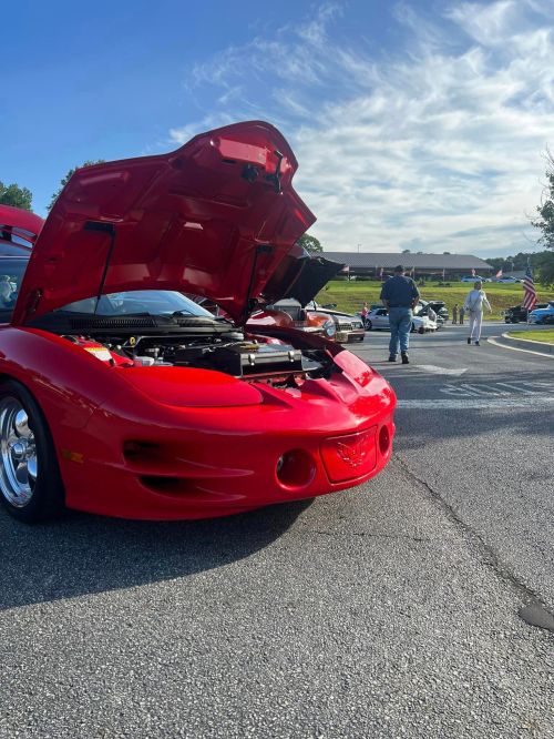 A red sports car with its hood open, parked at a car show with people and other vehicles in the background.
