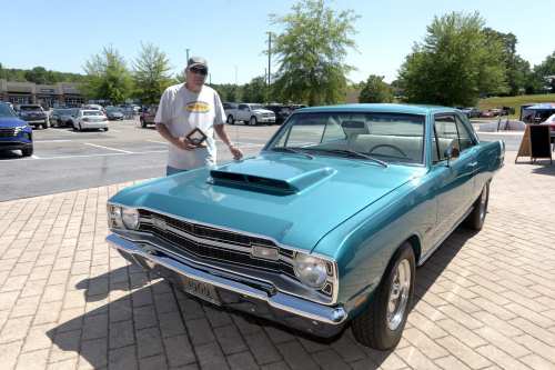 A man stands next to a turquoise classic car with a hood scoop, holding a trophy in a parking lot.