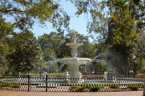 A decorative white fountain surrounded by lush greenery and trees, with water spraying from its tiers.
