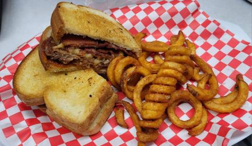 A grilled sandwich with layers of meat and cheese, served with curly fries on a red and white checkered paper.