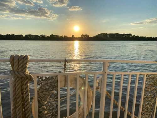 Sunset over a calm river, with a boat railing and a life ring in the foreground.