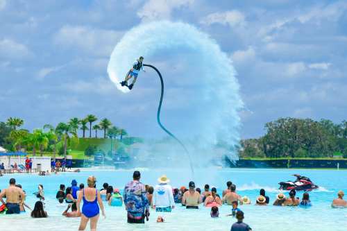 A person performs a water jetpack stunt above a crowd at a sunny beach, with palm trees in the background.