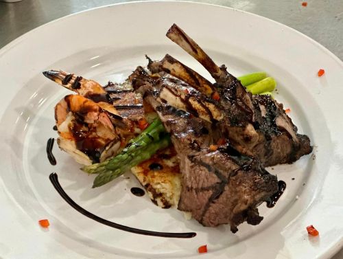 A gourmet plate featuring grilled lamb chops, shrimp, asparagus, and a drizzle of balsamic reduction on a bed of bread.