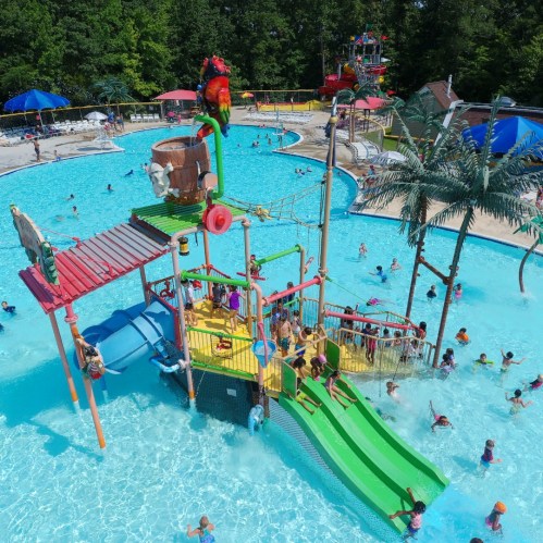 A vibrant water park with a large play structure, slides, and many people enjoying the pool on a sunny day.