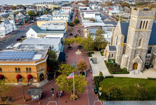 Aerial view of a charming town with brick streets, shops, and a church, featuring trees and an American flag.