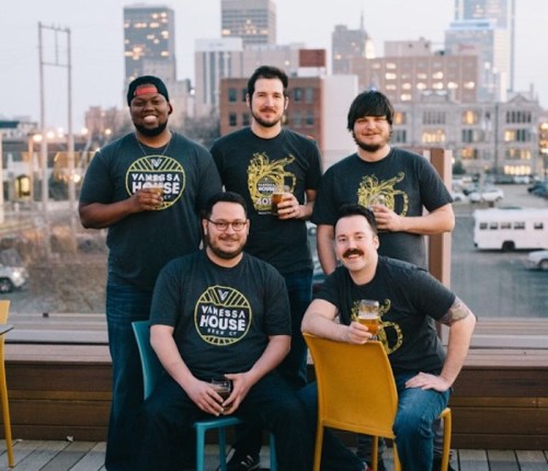 Five men in matching shirts pose with beers on a rooftop, city skyline in the background during sunset.