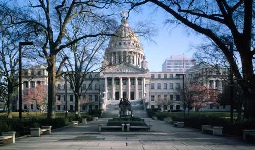 A grand government building with a dome, surrounded by trees and a statue in the foreground, under a clear blue sky.