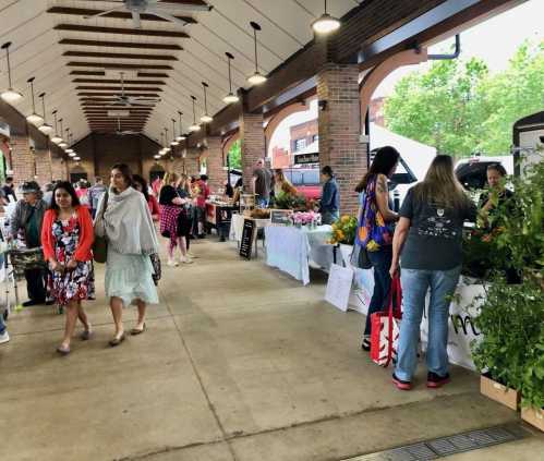 A bustling market scene with people browsing stalls filled with flowers and produce under a covered area.