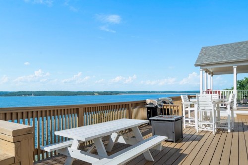 A scenic deck overlooking a calm lake, featuring picnic tables, grills, and a clear blue sky.