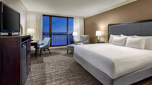 A modern hotel room featuring a king bed, seating area, desk, and large window with a view of the water.