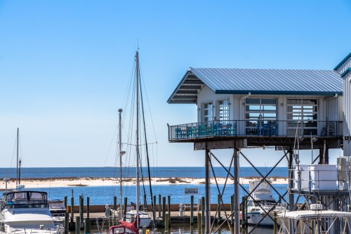 A coastal scene featuring a marina with boats and a stilted building overlooking the ocean under a clear blue sky.