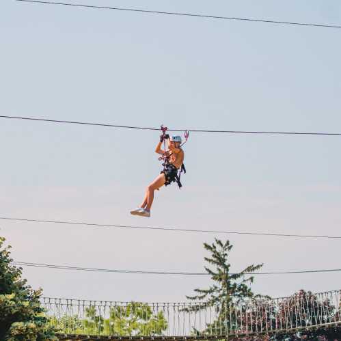 A person zip-lining across a clear blue sky, surrounded by trees and a scenic landscape below.