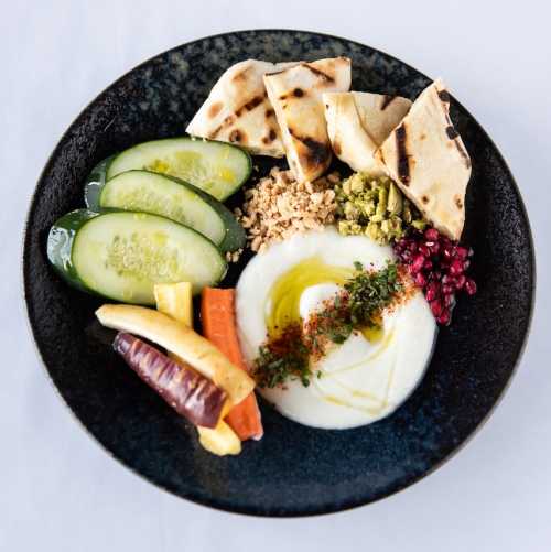 A plate featuring yogurt, cucumbers, carrots, pita, nuts, and pomegranate, garnished with herbs and olive oil.