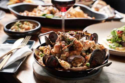 A close-up of a seafood dish with shrimp, clams, and herbs, surrounded by various side dishes and a glass of red wine.