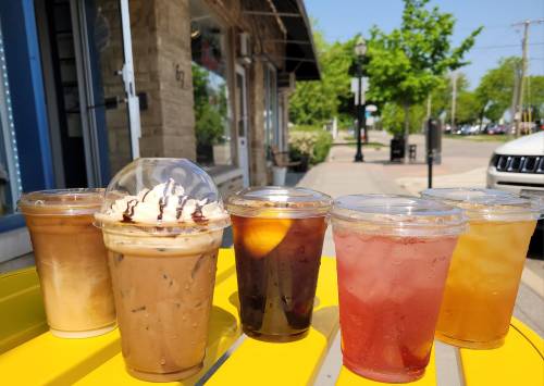 A row of six colorful drinks in clear cups on a sunny street, featuring iced coffee and fruity beverages.