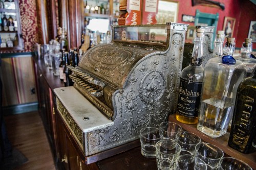 An ornate vintage cash register sits on a bar, surrounded by bottles and glasses in a warmly decorated setting.