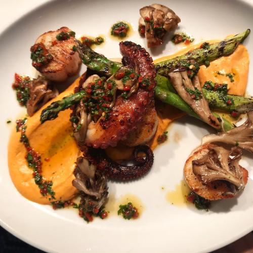 A gourmet dish featuring grilled octopus, scallops, asparagus, and mushrooms on a vibrant orange puree, garnished with herbs.