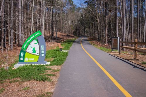A paved trail through a wooded area, with a trail map sign and a wooden fence on the side.