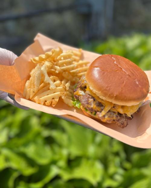 A hand holds a tray with a cheeseburger and crinkle-cut fries, set against a backdrop of fresh green lettuce.