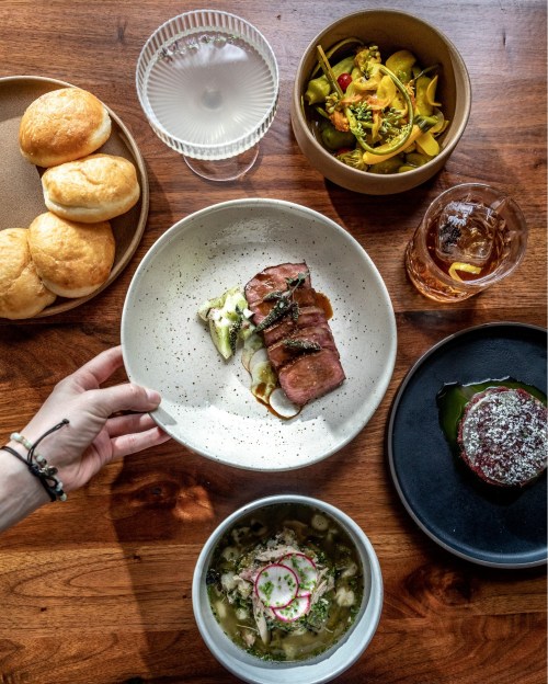 A hand reaches for a plate of sliced meat, surrounded by various dishes, rolls, and drinks on a wooden table.