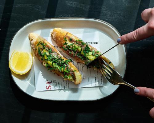 A plate with two stuffed bread rolls topped with herbs, accompanied by a lemon wedge and a hand holding a knife and fork.