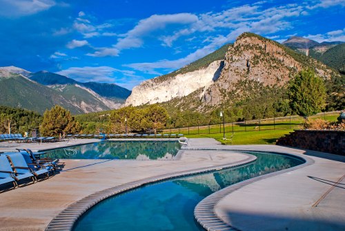 A scenic outdoor pool with a winding edge, surrounded by lush greenery and mountains under a bright blue sky.