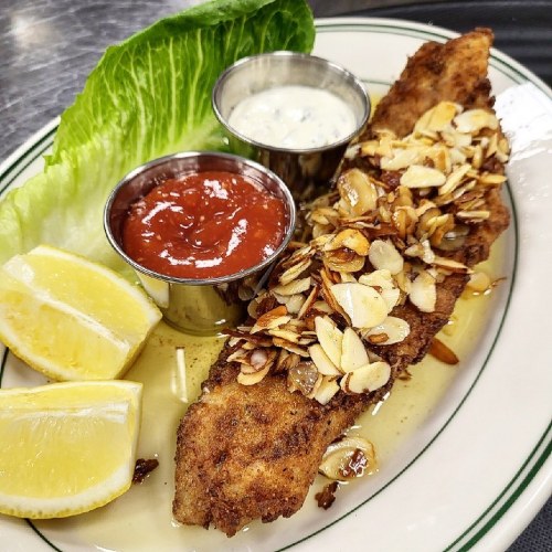 A breaded fish fillet topped with sliced almonds, served with lemon wedges, dipping sauces, and a lettuce leaf.