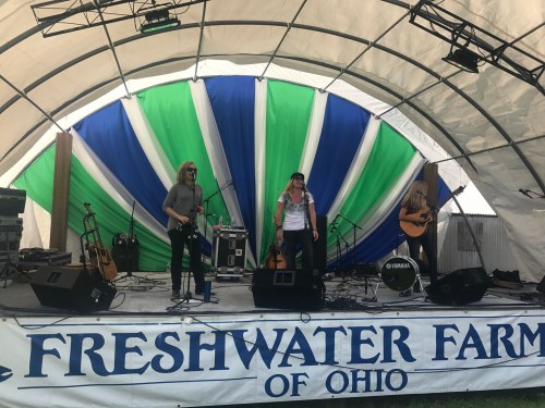 A band performs on stage at Freshwater Farms of Ohio, with colorful drapes and musical instruments visible.