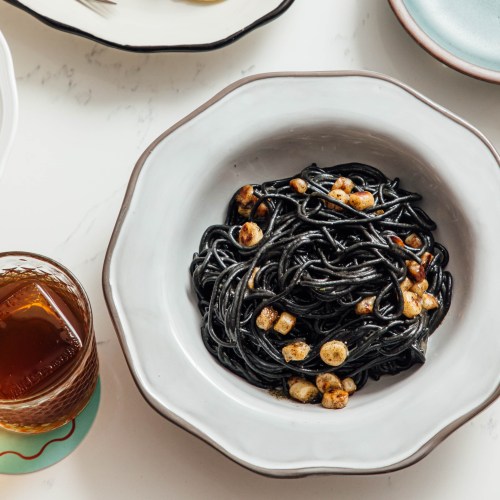 A bowl of black spaghetti topped with crispy bits, accompanied by a glass of iced drink on a marble surface.
