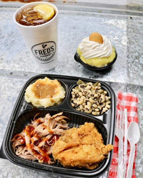 A takeout meal featuring fried chicken, pulled pork, mashed potatoes, black-eyed peas, and dessert with iced tea.