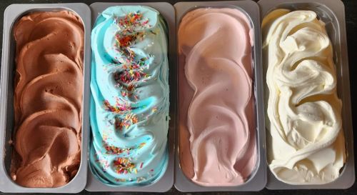 Four tubs of gelato: chocolate, blue with sprinkles, pink, and vanilla, all swirled and ready to serve.