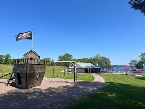 A pirate ship playground with a flag, near a lake and a building with outdoor seating under a clear blue sky.