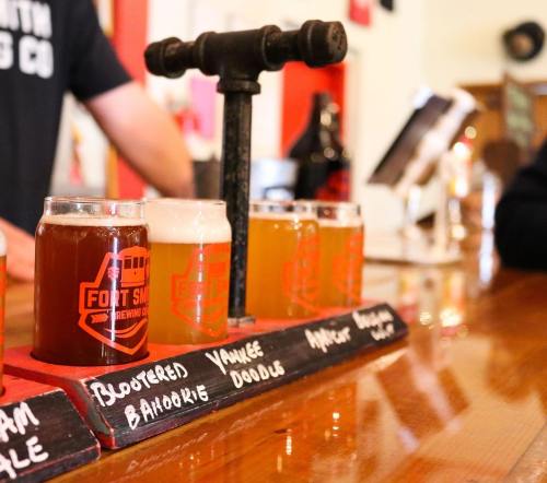 A wooden flight paddle with four small beer glasses in a brewery, featuring various beer styles and a tap in the background.